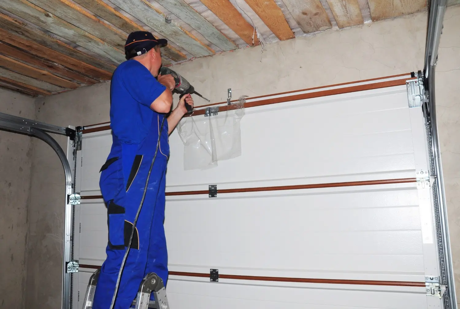 A technician on a ladder uses a power drill to install a garage door opener system on a white sectional garage door.
