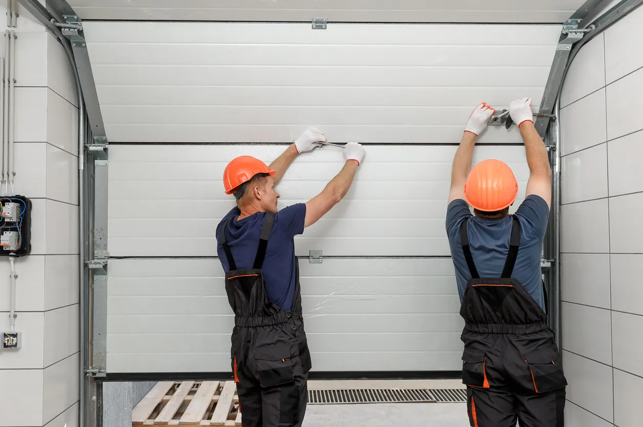 Two workers wearing orange hard hats and dark overalls are standing on a platform, using tools to install a white sectional garage door in a tiled workshop or garage.